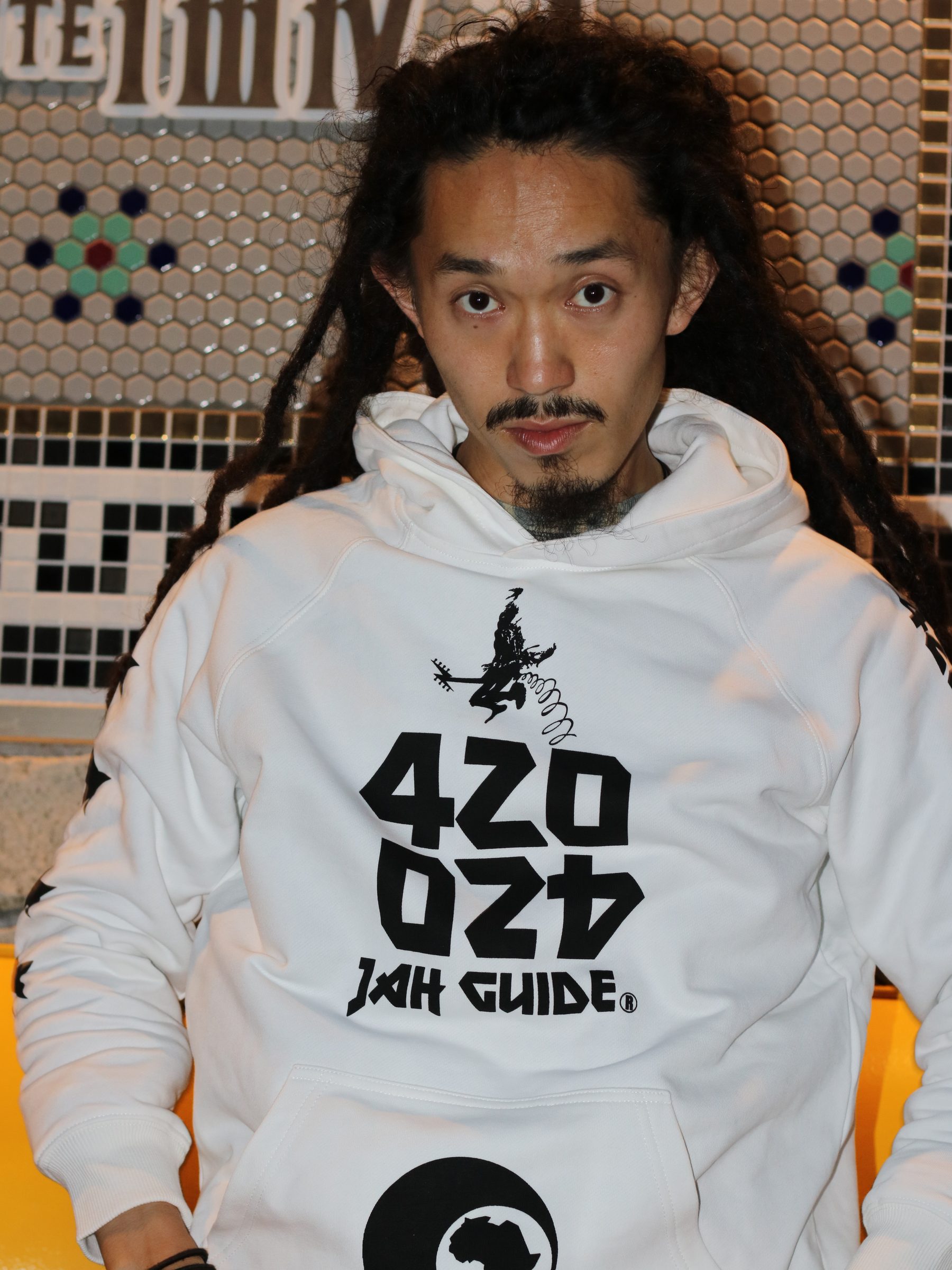 A man wearing the white JAH GUIDE logo hoodie, seated against a tiled wall